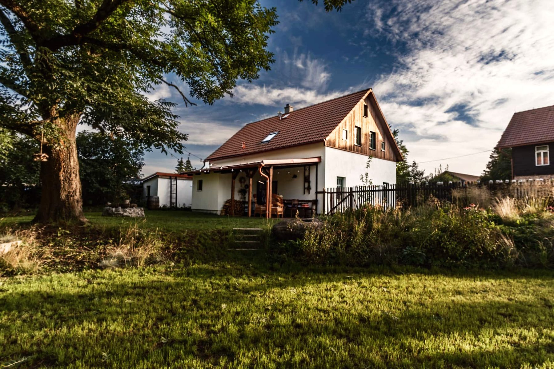 Bílé stavení se sedlovou střechou a dřevěným štítem, vlevo mohutný strom, před domem louka